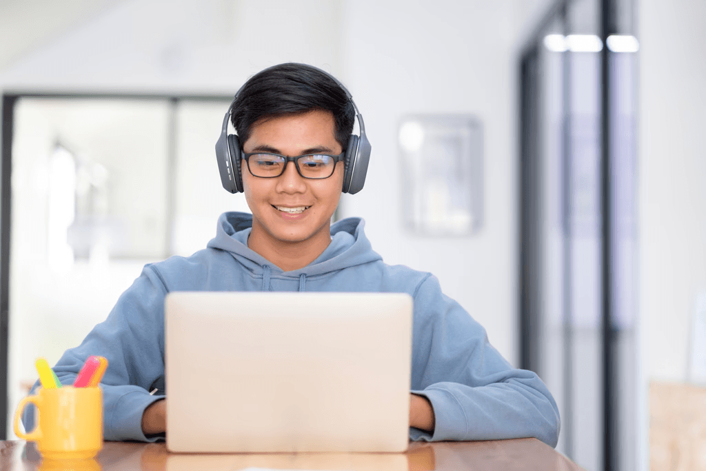 Smiling student wearing headphones, working on a laptop at a wooden desk in a bright indoor space