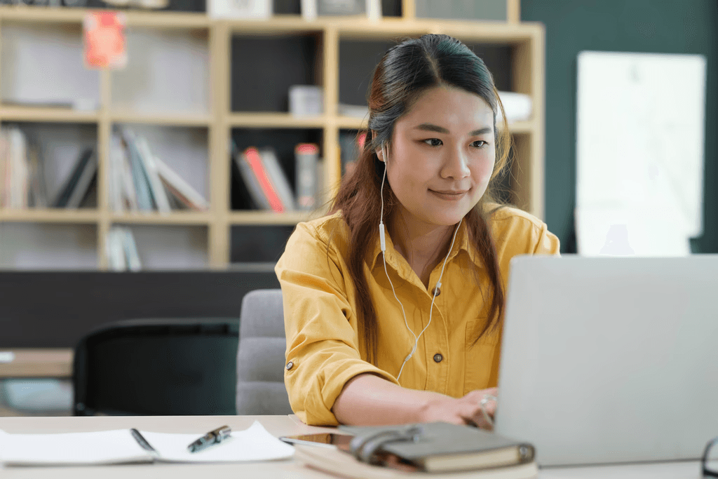 Student with earphones using a laptop beside a notebook and phone, with a bookshelf in the background