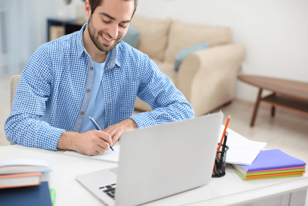 Writer in a checkered shirt taking notes in a notebook at a desk with a laptop and bright stationery