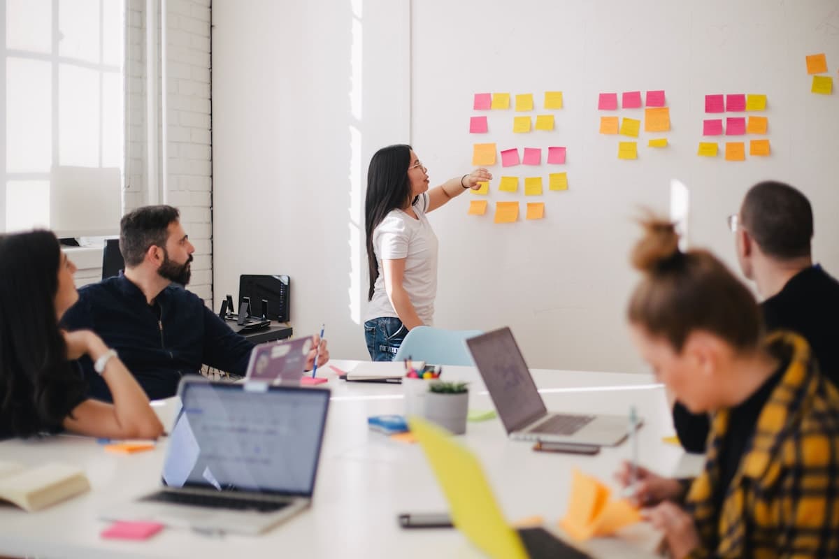 Workshop with sticky notes on a wall, presenter pointing while teammates work at laptops at a long table