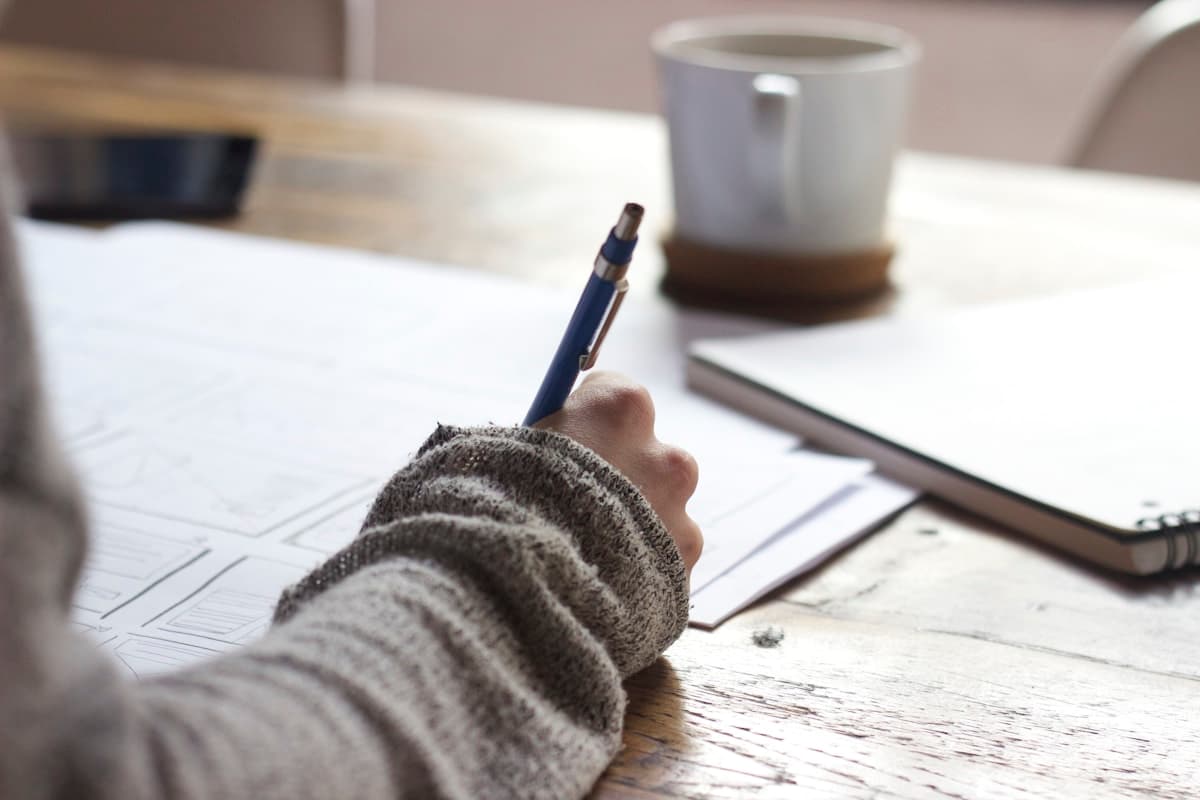 Hand with a blue pen drawing boxes and section lines on paper at a wood desk, coffee mug in soft focus